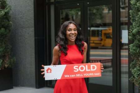 Smiling woman in red dress holding sold sign outside an office building.